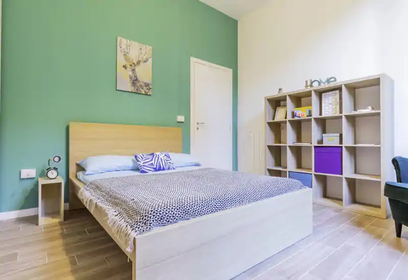 Camera da letto moderna e luminosa con grande letto in legno, biancheria a motivo e un mobile a cubi decorativo—parete accent color turchese e arredi essenziali creano un ambiente accogliente.