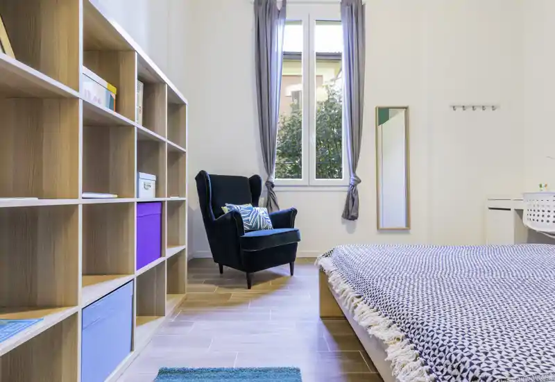 Camera da letto moderna e luminosa con letto basso in legno, plaid a motivi e una poltrona smeraldo vicino alla finestra; scaffalatura pulita per lo stoccaggio.