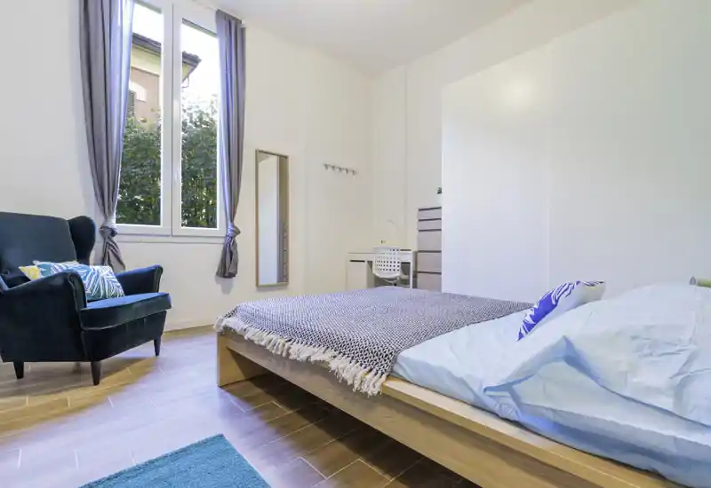 Camera da letto moderna e luminosa con letto su pedana in legno, tessili accoglienti e una poltrona comoda vicino alla finestra che lascia entrare luce naturale.