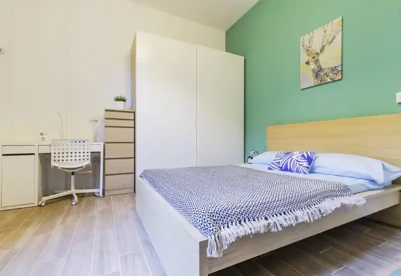 Camera da letto moderna e luminosa con letto in legno completo, coperta decorativa e parete accent color turchese; armadio e scrivania piccola inclusi, ideale per una persona o una coppia.