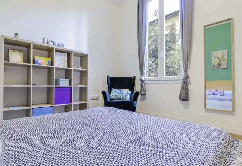 Camera da letto moderna e luminosa con copriletto a fantasia, una poltrona accogliente e scaffalatura per riporre oggetti. Ampia finestra che dona molta luce naturale e pareti neutre per un’atmosfera rilassante.