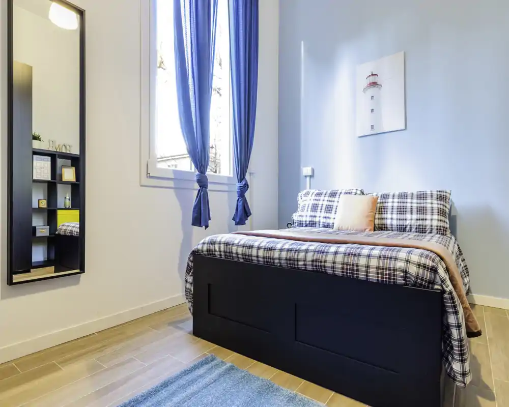 Dormitorio luminoso y ordenado con cama completa, ropa de cama a cuadros y cortinas azules a juego. Ventana grande que aporta luz natural, suelo de madera claro y espejo de cuerpo entero.