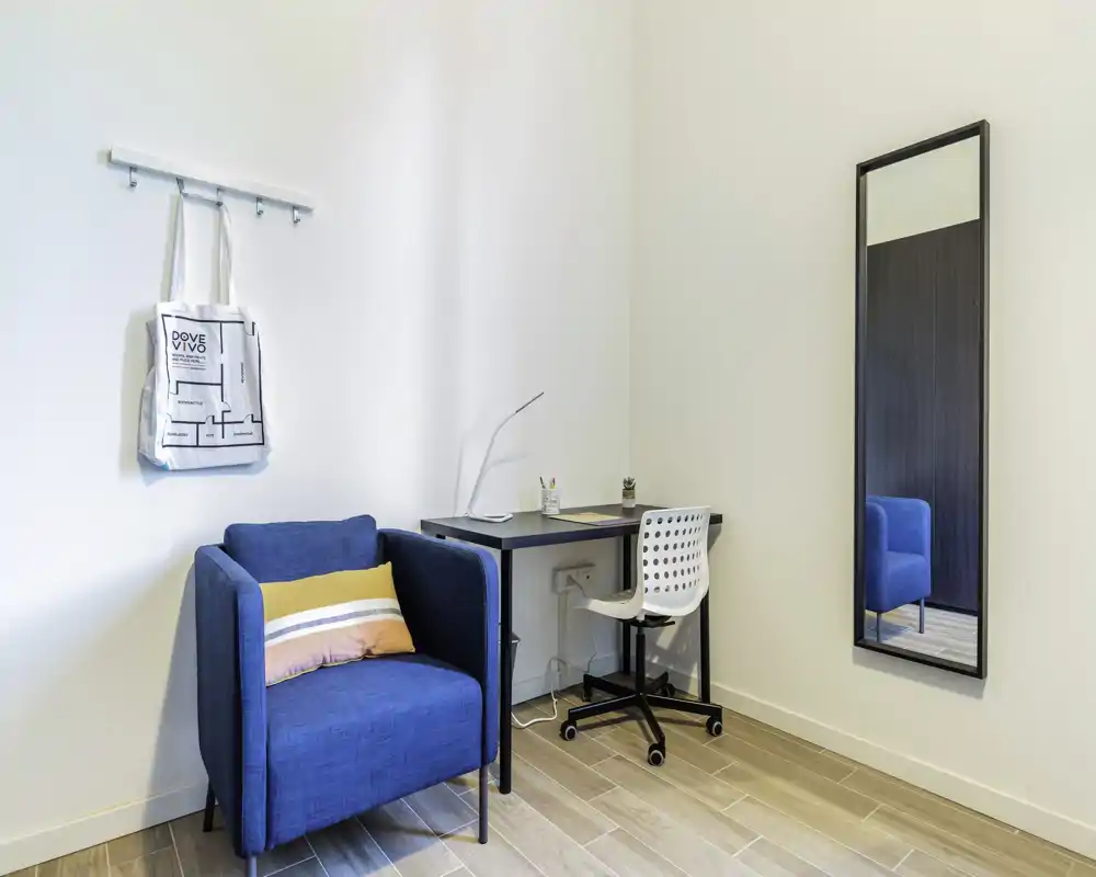 Rincón de estar moderno y luminoso con un sillón azul, escritorio compacto y espejo de cuerpo entero: un espacio ordenado y bien iluminado, perfecto para descansar o trabajar.