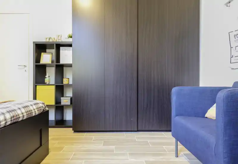 Rincón de dormitorio luminoso y moderno con cama parcialmente visible, gran armario oscuro y un sillón azul; suelo de madera clara y líneas limpias que aportan una sensación contemporánea.