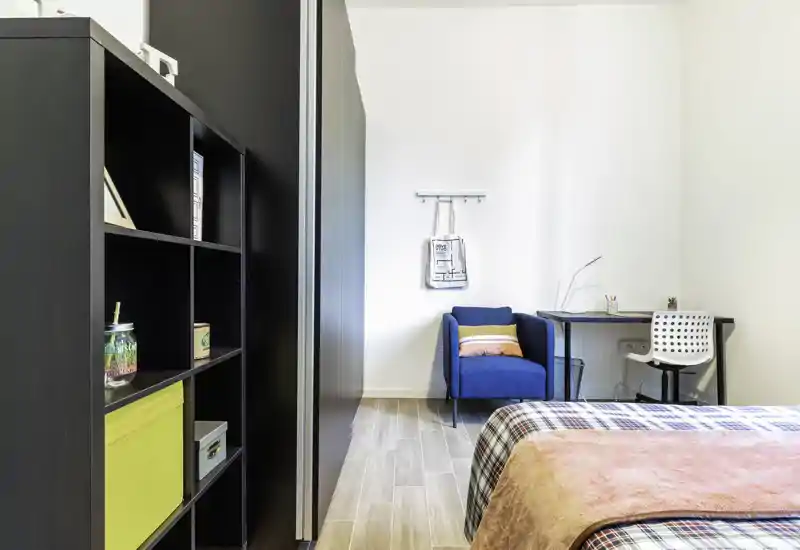 Dormitorio moderno y luminoso con cama visible, manta cálida, butaca azul y pequeño escritorio: ambiente limpio y acogedor perfecto para alquileres temporales.