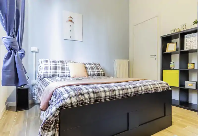 Dormitorio luminoso y bien presentado con una cama grande de cuadros, manta y cojín suaves y una estantería para almacenaje — perfecto para anuncios que destaquen comodidad y luz natural.