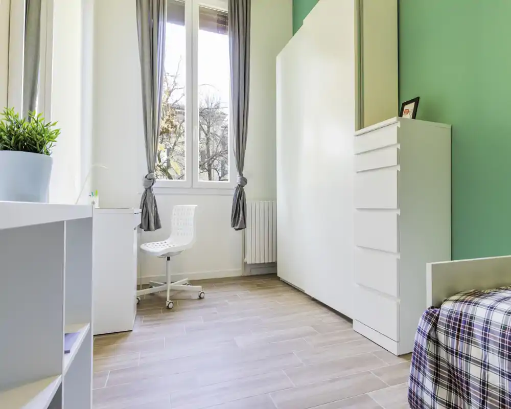Bright, simply staged bedroom corner with a single bed partially visible, white storage chest and a desk by a large window letting in natural light.