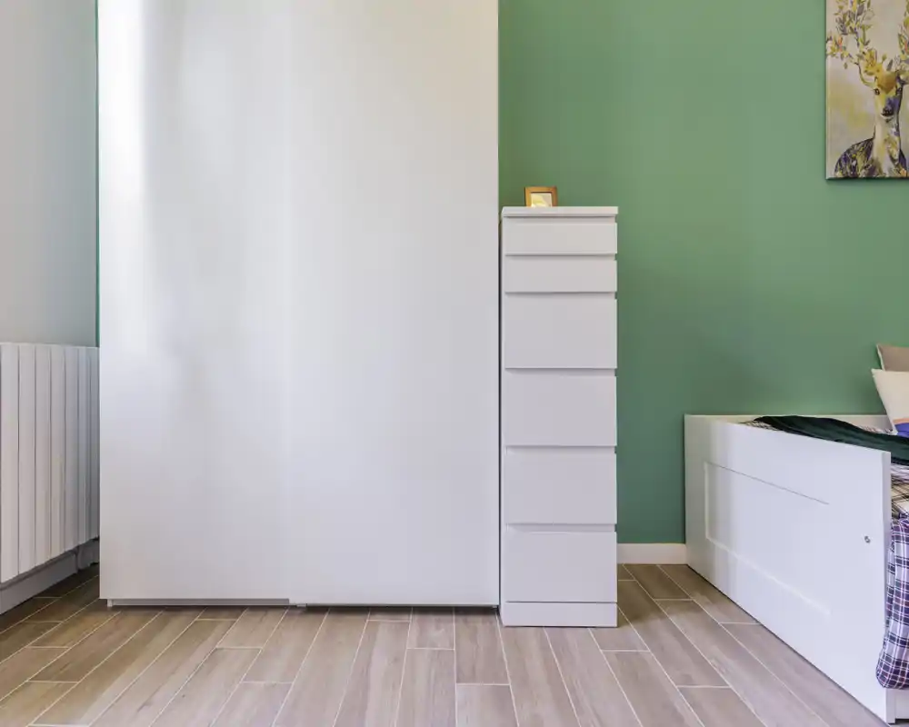 Bright, minimal bedroom corner with white furniture and a green accent wall; partial view of a single bed and a tall chest of drawers, ideal for compact listings.