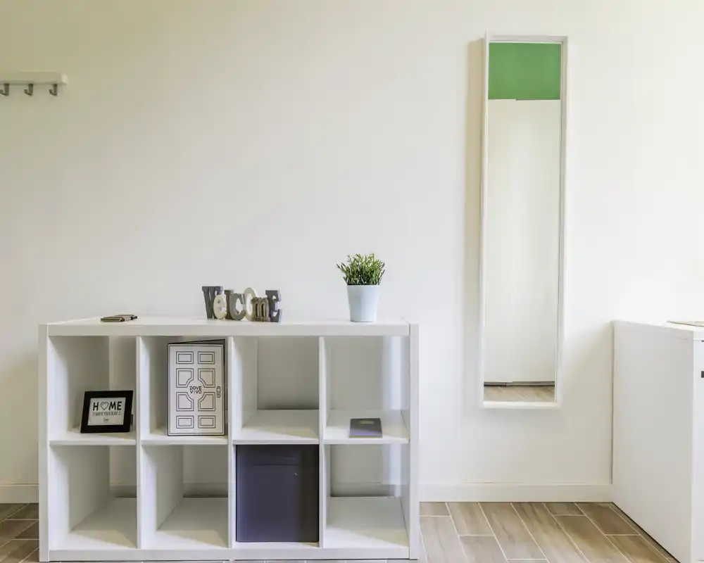 Minimal, bright entrance area with a white cubby shelf, decorative items and a tall mirror — clean, modern welcome space.