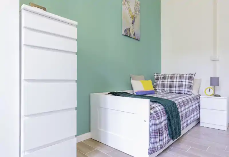 Compact, well-styled single bedroom with a white bed frame, plaid bedding and a cozy teal throw against a mint-green accent wall. Bright, clean and ready for a comfortable stay.