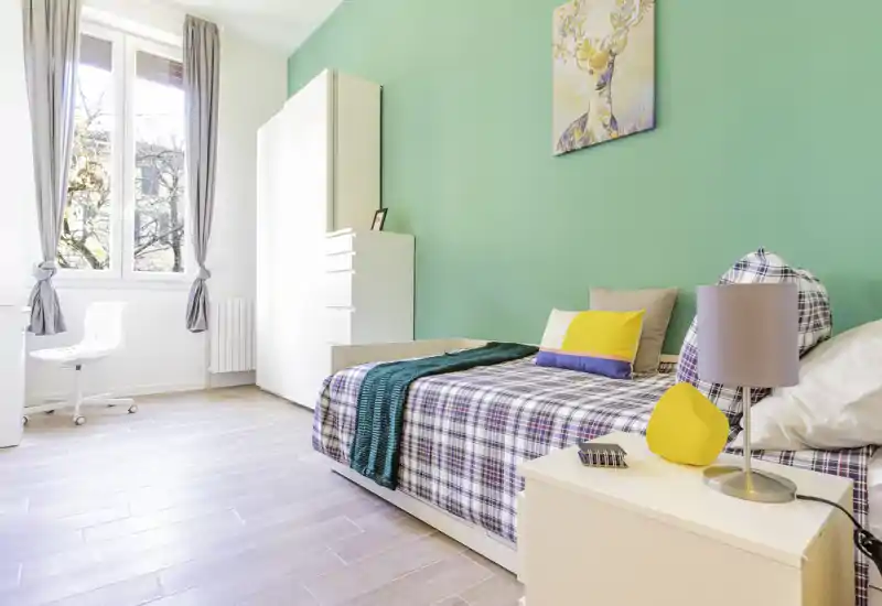 Bright, modern single bedroom with a plaid bed, colorful cushions and a mint-green accent wall; large window provides ample natural light and a tidy wardrobe for storage.