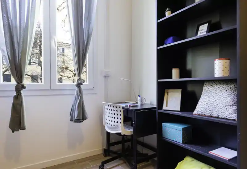 Bright small living/study corner with a desk by a large window and open shelving. Natural light and tidy decor create a pleasant, versatile space suitable for work or relaxation.