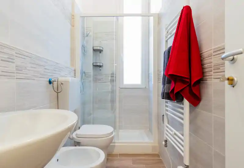 Bagno moderno e pulito con doccia a vetri, WC, bidet e lavabo a parete. Piastrelle neutre e termosifone portasciugamani con asciugamani colorati donano un tocco fresco e accogliente.