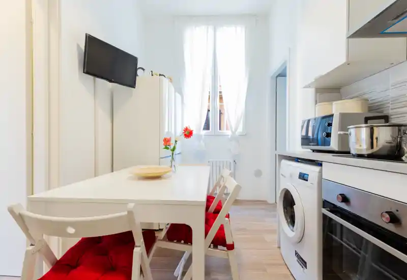 Bright, compact eat-in kitchen with white furnishings, a table with red-cushioned chairs, built-in appliances and a window providing natural light.