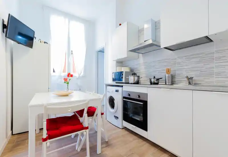 Bright modern kitchen with white cabinets, integrated oven, washing machine and a small dining table with red cushions — airy natural light from the window.