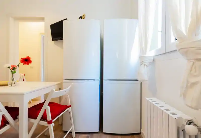 Bright, compact kitchen corner with two white refrigerators, a small dining table and red-cushioned folding chairs; airy window light and simple, modern styling.