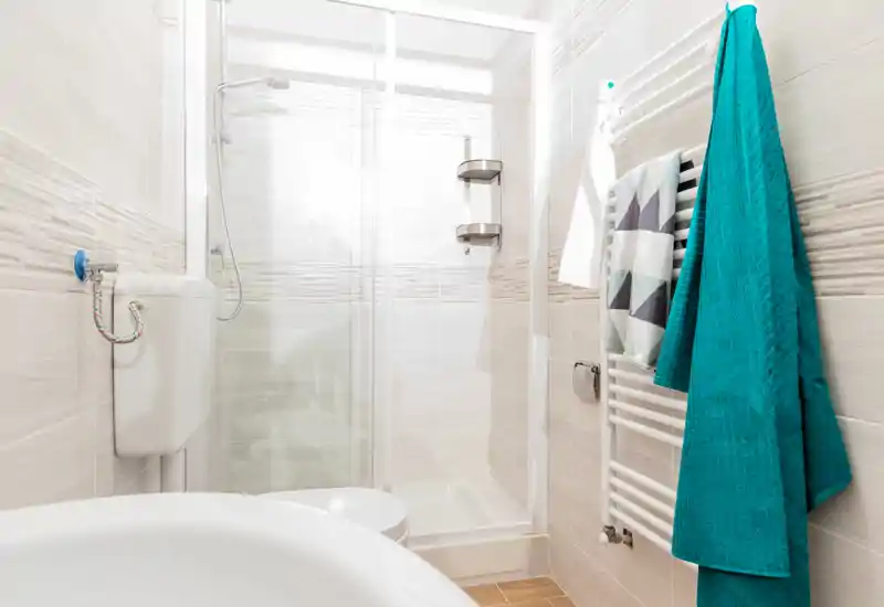 Bright, modern bathroom with a glass shower enclosure, wall-mounted radiator with towels, and compact fixtures in a light tile finish.