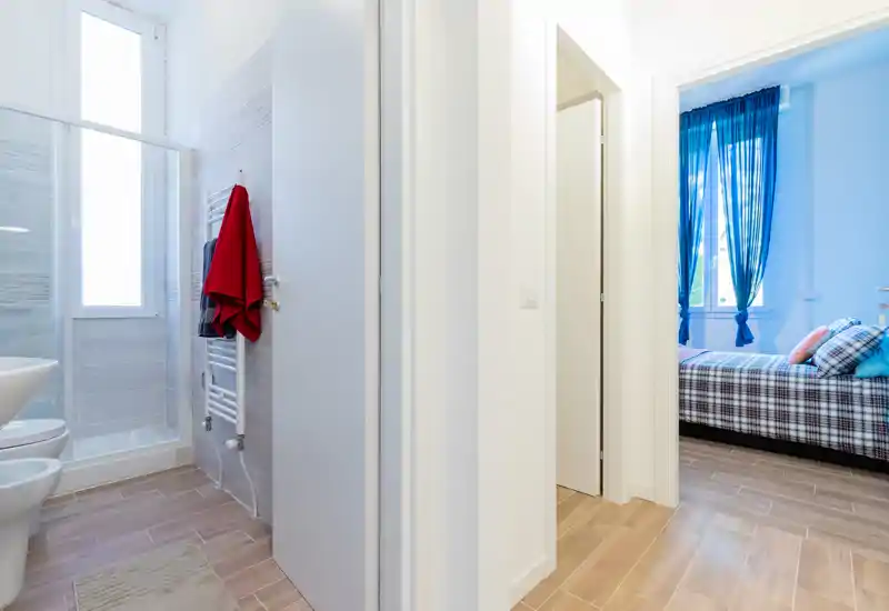 Bright, modern bedroom seen through a hallway: a neatly made single/double bed with patterned bedding and colorful cushions, framed by blue curtains and abundant natural light.