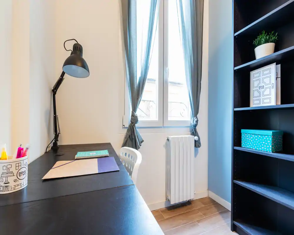 Angolo studio luminoso con scrivania nera, lampada regolabile, sedia e scaffale; la luce naturale dalla finestra con tende crea un ambiente accogliente per lavorare.
