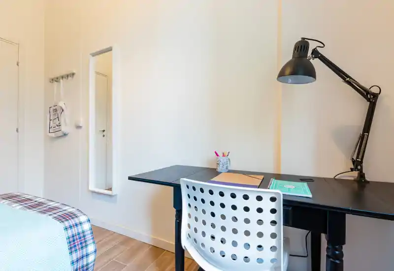 Bright, tidy bedroom corner with a study desk. The image shows a neatly made bed at the left and a functional black desk with a lamp and chair, ideal for a student or home worker.