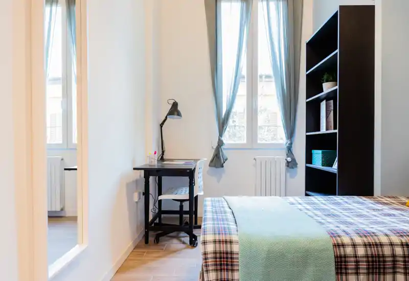 Bright, neatly styled bedroom with a comfortable double bed, a small study desk by the window, and an open shelving unit — ideal for a short-term rental or student flat.
