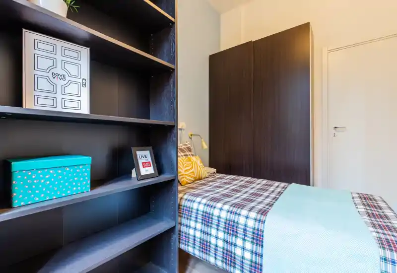Compact single bedroom with a patterned bedspread, decorative cushions and a tall dark wardrobe; shelving in the foreground provides storage and styling options.
