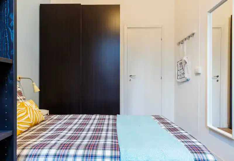 Bright, neatly styled bedroom with a large bed dressed in patterned linens, decorative pillows and a pastel throw, plus a dark wardrobe and full-length mirror that add depth.