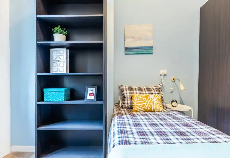 Bright, tidy single bedroom with a checked bedspread, decorative cushion, bedside lamp and shelving unit — ideal for a comfortable short-stay.