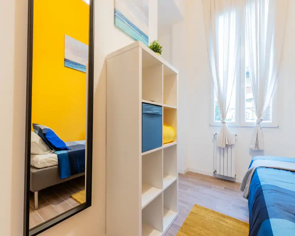 Chambre lumineuse et bien décorée avec lit soigneusement fait, rideaux clairs et étagère; touches jaunes et bleues apportent une ambiance chaleureuse.
