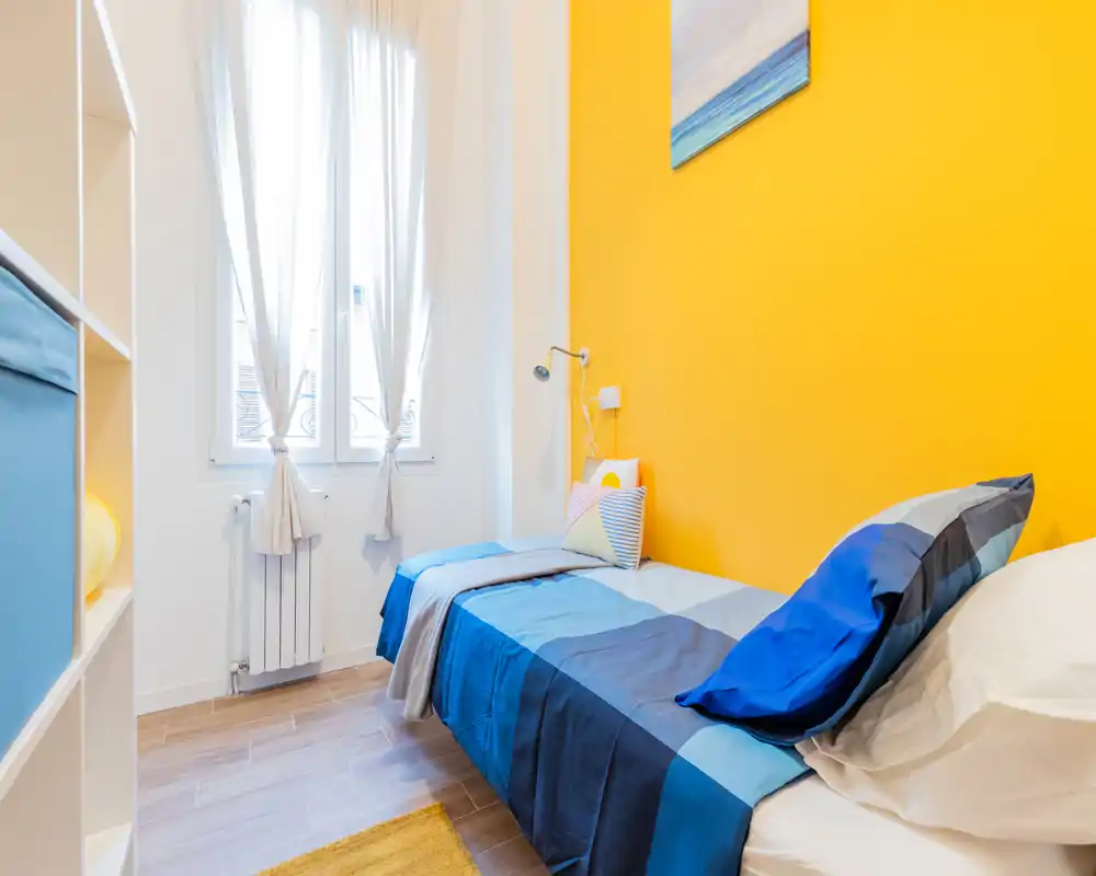 Chambre simple lumineuse avec une couette bleue sur un mur d'accent jaune. La lumière naturelle de la fenêtre met en valeur le linge de lit soigné, la lampe de lecture et les coussins décoratifs.