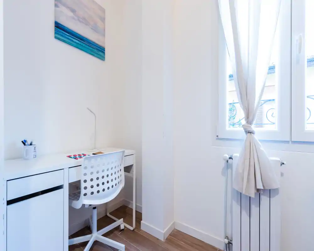 Coin bureau lumineux et minimaliste avec un bureau blanc, chaise pivotante perforée et lumière naturelle venant de la fenêtre — espace de travail propre et fonctionnel.