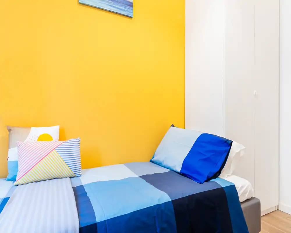 Chambre simple lumineuse avec literie bleue colorée, coussins décoratifs et un mur d'accent jaune vif. Espace propre et bien éclairé, idéal pour une annonce locative.