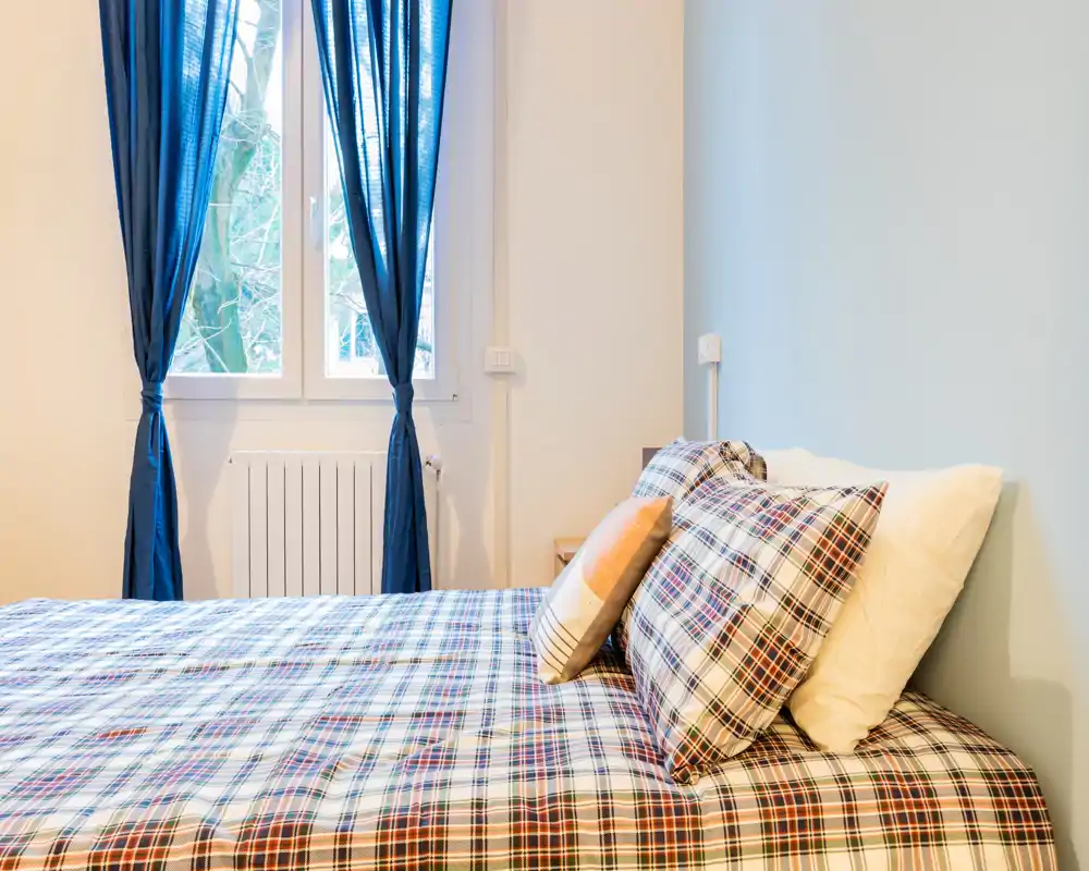 Dormitorio luminoso y bien decorado con una gran cama de cuadros en primer plano y luz natural procedente de una ventana con cortinas azules.