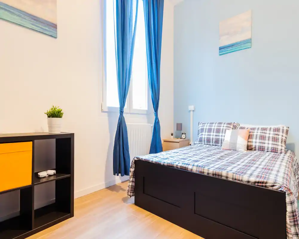 Dormitorio luminoso y ordenado con cama doble y ropa de cama a cuadros, luz natural desde la ventana con cortinas azules y mobiliario moderno sencillo.