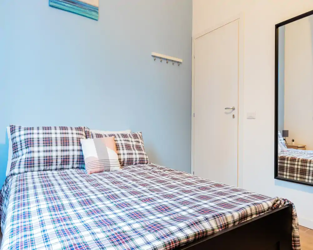 Dormitorio luminoso y ordenado con cama doble con ropa de cama a cuadros, cojines decorativos y un espejo de cuerpo entero en la pared — ideal para una estancia cómoda.