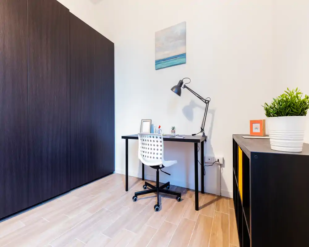 Rincón de trabajo luminoso y minimalista con escritorio negro, lámpara articulada, silla con ruedas y gran armario; líneas limpias y una planta que aporta verde.