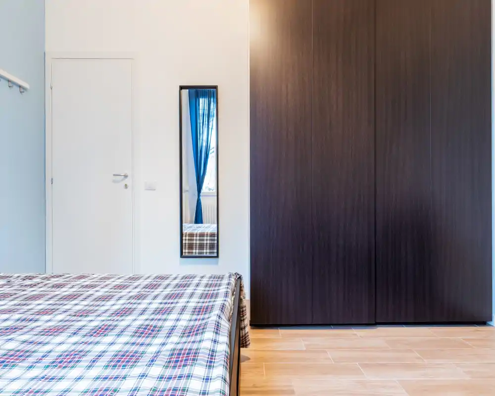 Dormitorio moderno y luminoso con cama con colcha a cuadros y un gran armario oscuro; líneas sencillas y luz natural que lo hacen práctico y acogedor.