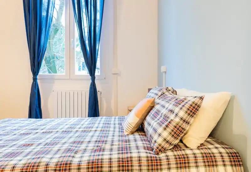Dormitorio luminoso y bien decorado con una gran cama de cuadros en primer plano y luz natural procedente de una ventana con cortinas azules.