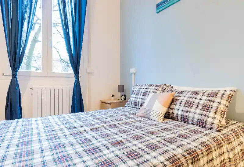 Dormitorio luminoso y bien arreglado con cama grande de cuadros, cojín decorativo y luz natural desde la ventana con cortinas azules — ideal para anuncios.