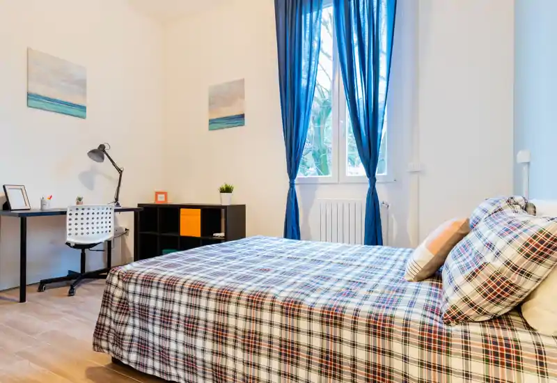 Dormitorio luminoso y bien presentado con una amplia cama de estampado, escritorio y luz natural desde la ventana con cortinas azules — acogedor y bien iluminado.