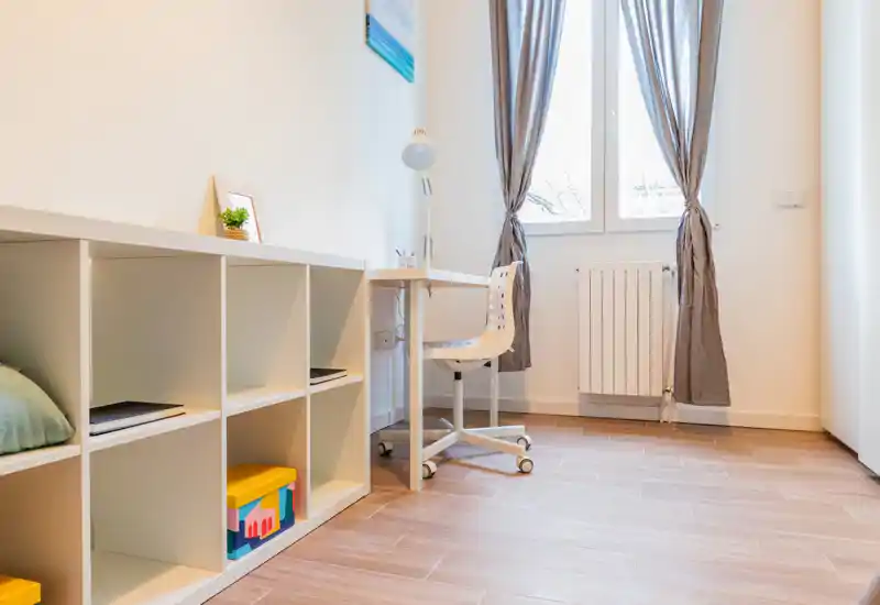Bright, minimal study corner with a white desk, rolling chair and modular shelving. Natural light from a window and simple decor create a clean, functional workspace.