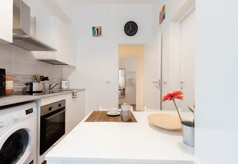 Bright, compact kitchen with modern appliances, a built-in oven and washing machine, and a small dining surface set with a cup and decorative flower.