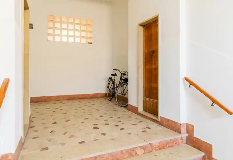 Bright, clean building entrance with tiled floor, wooden door and handrails; a bicycle stored against the wall and natural light through a glass-block window.