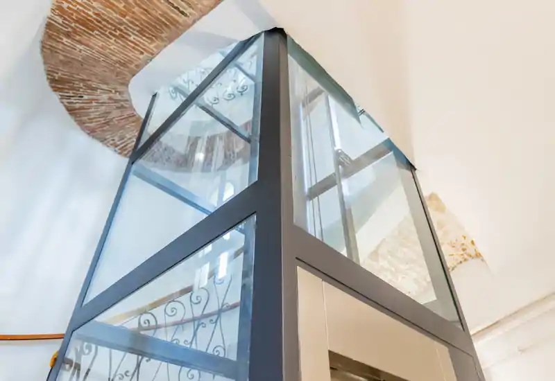 Modern glass elevator shaft inside a building entrance area, showing metal framing and decorative wrought-iron stair railing in the background.