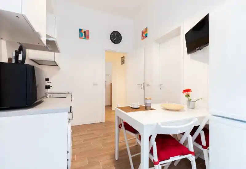 Compact modern kitchen with a small dining table, red-cushioned chairs, countertop appliances and bright white walls — ideal for a city apartment.