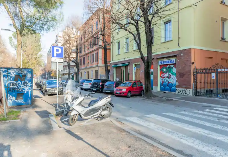 Sunny urban street scene with parked cars and scooters along a tree-lined sidewalk; storefronts and apartment buildings in the background.