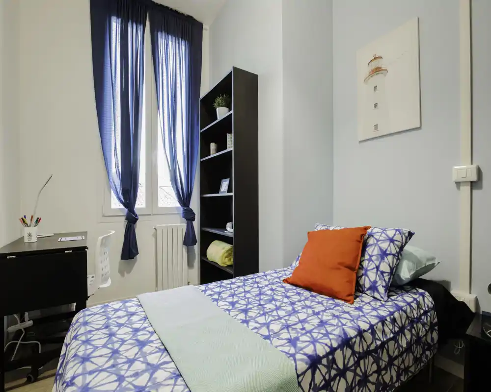 Camera singola luminosa e ordinata con biancheria fantasia, cuscini colorati e scrivania vicino alla finestra — perfetta per uno studente o un viaggiatore singolo.