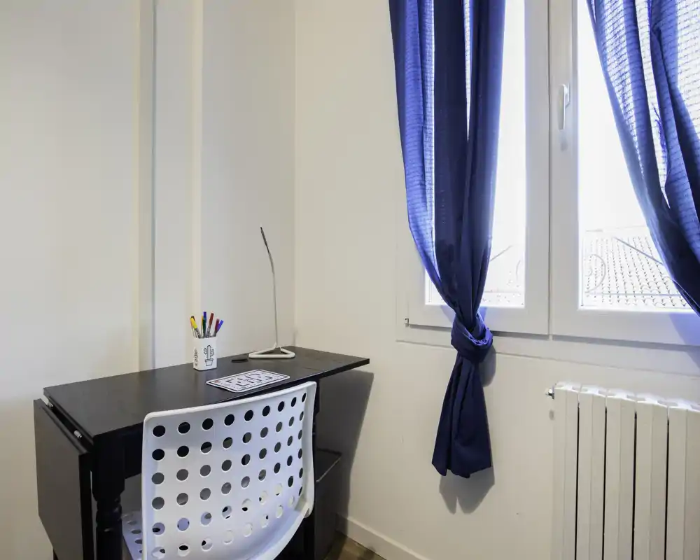 Luminoso angolo lavoro con scrivania nera moderna, sedia bianca forata e finestra con tende blu, ideale per studio o smart working.