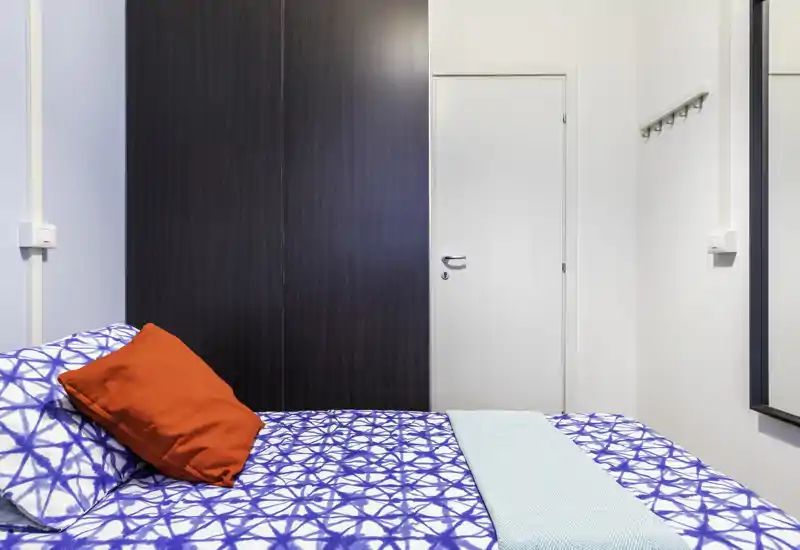 Camera da letto luminosa e ordinata con copriletto blu a motivi, cuscino rosso come elemento d'accento e armadio a muro. L'immagine mette in evidenza l'area del letto e un arredamento moderno e minimale.