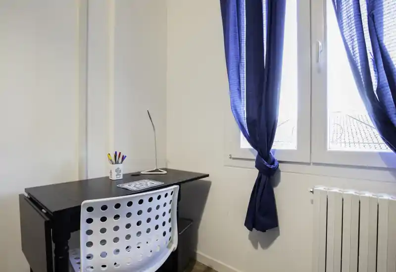 Luminoso angolo lavoro con scrivania nera moderna, sedia bianca forata e finestra con tende blu, ideale per studio o smart working.